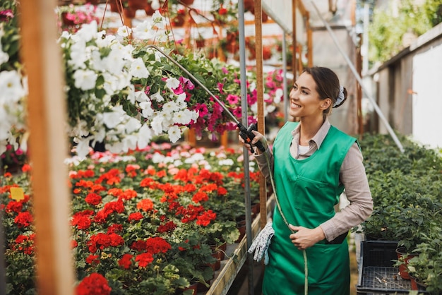 Eine lächelnde Frau in grüner Schürze, die Blumen in einem Gartencenter oder einer Gärtnerei gießt.