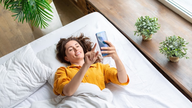 Eine lächelnde Frau liegt nach dem Aufwachen mit Handy im Bett