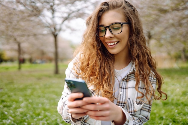 Eine lächelnde Frau mit Brille hält ein Handy, während sie auf einer grünen Wiese in einem Blumenpark sitzt