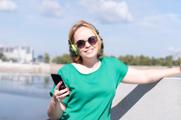 Eine lächelnde Frau mit Kopfhörern, die ein Telefon in der Hand hält, hört Musik, die am Flussufer steht