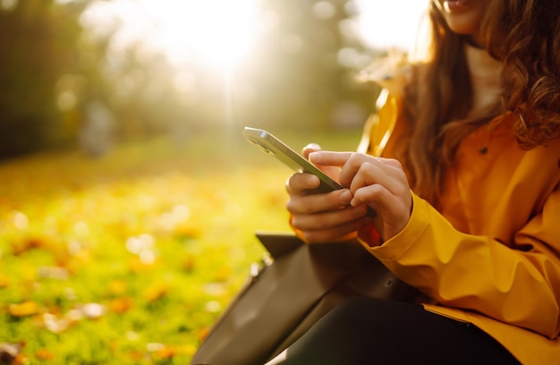 Eine lächelnde Frau sitzt draußen auf dem Rasen inmitten gelber Herbstblätter und hält ein Telefon in der Hand