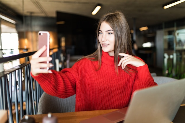 Eine lächelnde Freelancer-Frau macht ein Selfie am Telefon oder macht einen Videoanruf, einen Arbeitstag einer freien jungen Frau oder eines Studenten mit einem Laptop.