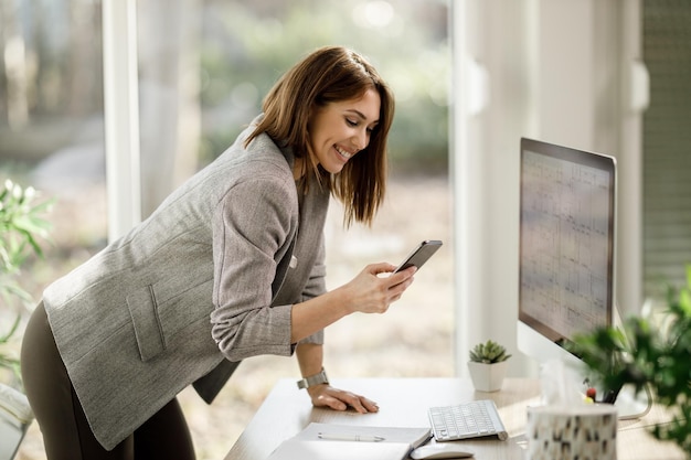 Eine lächelnde Geschäftsfrau, die Smartphone benutzt, während sie in ihrem Heimbüro am Computer arbeitet.