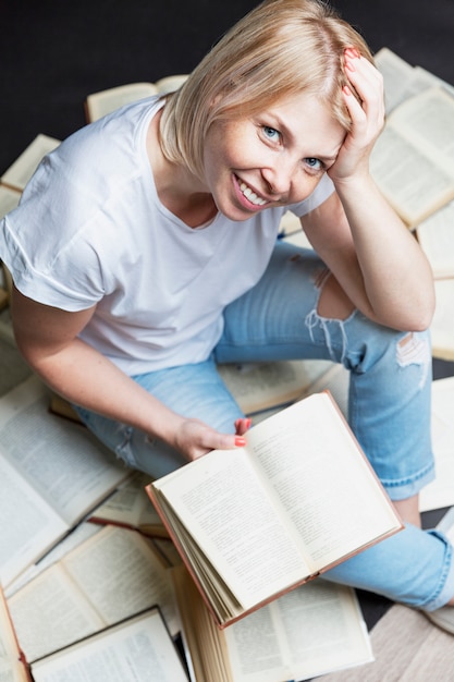 Eine lächelnde junge blonde Frau sitzt mit einem Stapel Bücher und liest. Schul-und Berufsbildung. Vertikale.