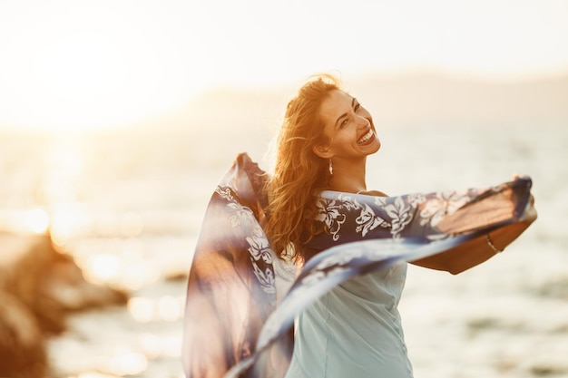 Eine lächelnde junge Frau amüsiert sich und genießt einen Sommerurlaub. Sie posiert mit einem Schal in den Händen am Strand.