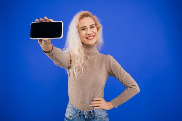 Eine lächelnde junge Frau hält einen Telefonbildschirm an die Kamera auf einem blauen Hintergrund Mockup