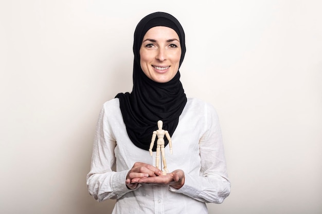 Eine lächelnde junge Frau in einem Hijab hält eine Marionette eines hölzernen Mannes auf hellem Hintergrund