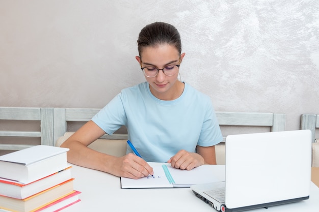 Eine lächelnde Studentin, die mit einem Laptop an einem Tisch sitzt, schreibt eine Aufgabe in ein Notizbuch. Studentin studiert zu Hause. Zurück zum Schulkonzept. Schulkonzept zu Hause.