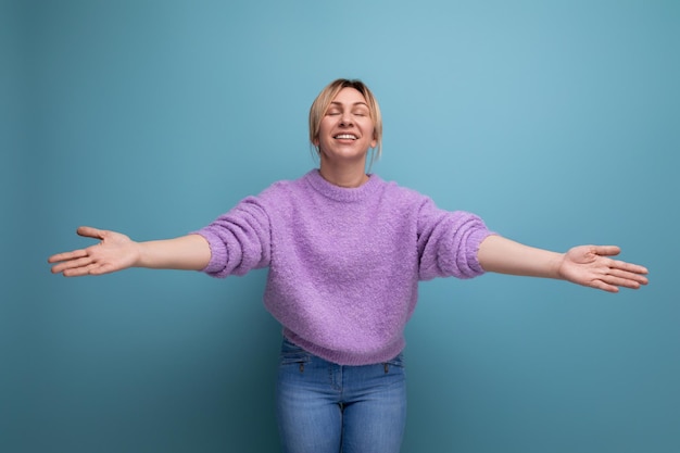 Foto eine lächelnde, süße blonde junge frau in einem fliederfarbenen pullover zeigt auf einem hellen hintergrund, wie sie sich zum umarmen ausstreckt