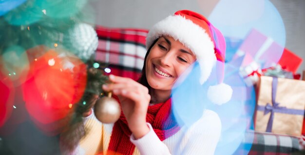 Eine lächelnde, wunderschöne junge Frau mit Weihnachtsmütze schmückt zu Hause einen großen Weihnachtsbaum und hat Spaß