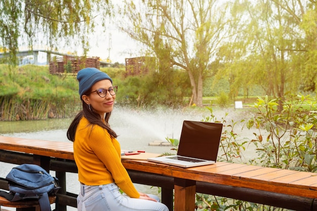 Eine lateinamerikanische Studentin sitzt in einem Park mit See und trägt eine blaue Mütze und einen gelben Pullover, während sie ihren Laptop benutzt und direkt in die Kamera schaut