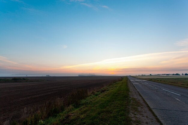 Eine leere Autobahn-Asphaltstraße durch die Felder und den Wald in einem dichten Nebel bei Sonnenaufgang. Stimmungsvolle Landschaft. Idyllische ländliche Szene. Dunkelheit, Herbstsaison, wechselhaftes Wetter, gefährliches Fahren, Roadtrip