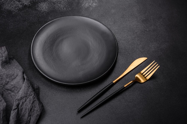 Eine leere schwarze Platte auf dunklem Betongrund mit Gewürzkräutern und Besteck