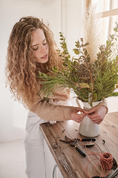 Eine lockige blonde Dame legt ein Seil um eine Vase mit kreativer Komposition, während sie während der Arbeit am Holztisch im Blumenstudio steht