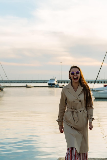 Eine lustige langhaarige Frau mit Sonnenbrille und beigem Mantel wartet bei Sonnenuntergang lachend auf ihr Boot im Seehafen