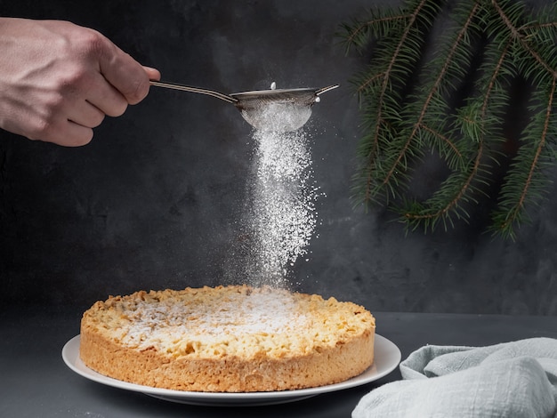 Eine männliche Hand streut Puderzucker auf einen Apfelkuchen, der auf einem Tisch steht.