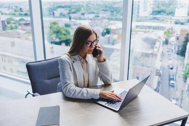 Eine Managerin mit Brille sitzt an einem Laptop in einem Büro mit atemberaubendem Blick auf die Stadt und führt ein Geschäftsgespräch am Telefon Geschäftsfrau, die am Holztisch im Büro arbeitet