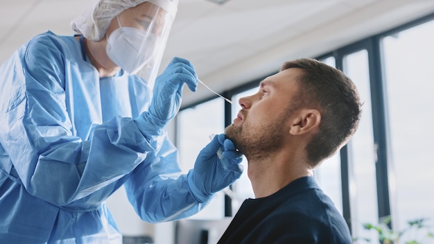 Eine medizinische Krankenschwester in Sicherheitshandschuhen und Maske, ein schützendes Gesichtsschild und ein Overall nehmen eine PCR-Coronavirus-Probe in einer Gesundheitsklinik.