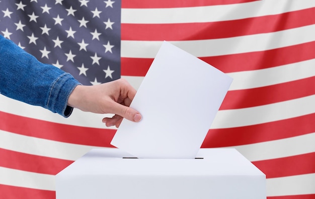 Eine menschliche Hand wirft einen Stimmzettel in die Wahlurne. Amerikanische Flagge im Hintergrund. Wahlkonzept