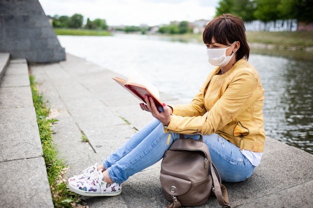 Eine mittelgroße Frau in Freizeitkleidung und medizinischer Maske sitzt an der Promenade und liest an einem sonnigen, warmen Frühlingstag ein Buch