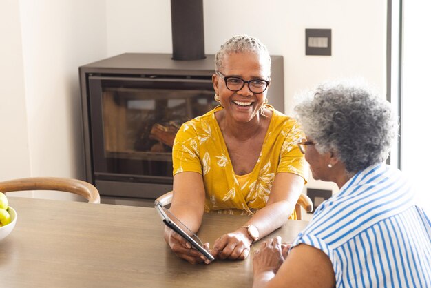 Eine mittelrassische ältere Frau in einem gelben Oberteil lacht zu Hause mit einer afroamerikanischen Frau