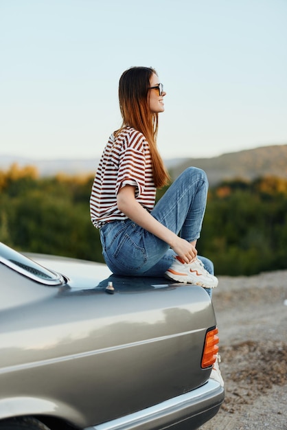 Eine Modefrau mit stylischer Brille, gestreiftem T-Shirt und Jeans sitzt auf dem Kofferraum eines Autos und blickt auf die wunderschöne Natur des Herbstes