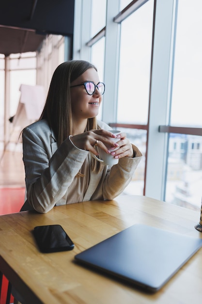 Eine moderne Frau mit slawischem Aussehen, eine Managerin in einer leichten Jacke und einer Brille, ein Mädchen mit einem Lächeln im Gesicht, sitzt an einem Tisch in einem Café und trinkt Tee Fernarbeit