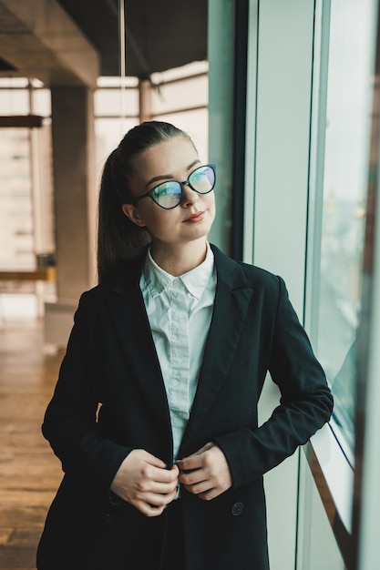 Foto eine moderne lächelnde geschäftsfrau in einem schwarzen anzug und brille steht im büro in der nähe des fensters frau manager in brille und weißem hemd