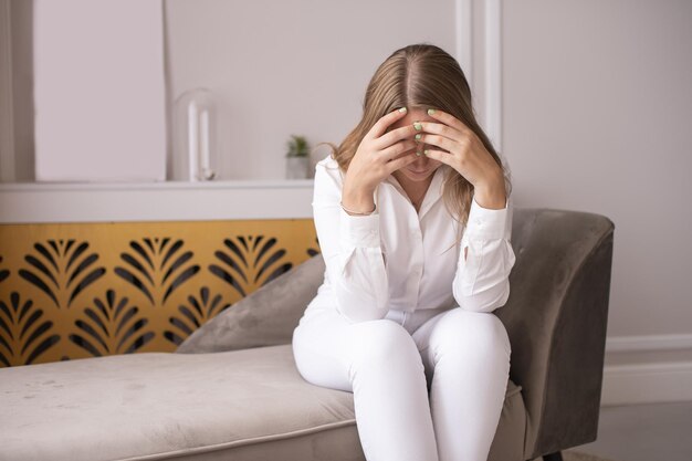 Eine müde Frau sitzt nach der Arbeit auf der Couch und schließt mit den Händen die Augen