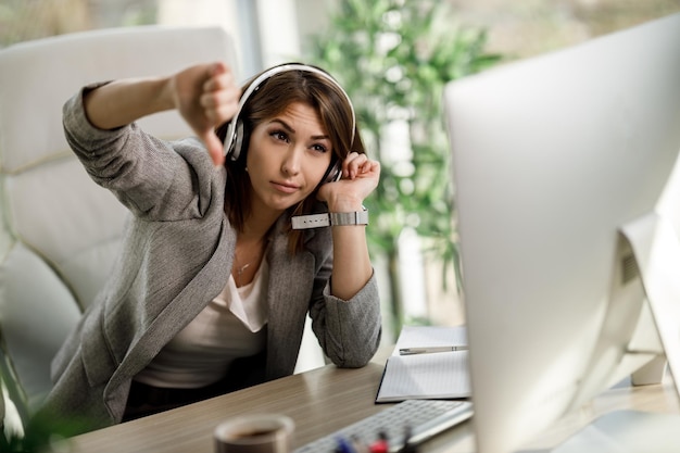 Eine müde junge Frau mit Kopfhörern, die während eines Videoanrufs in einem modernen Büro den Daumen nach unten zeigt.