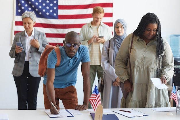 Eine multiethnische Gruppe von Personen, die sich am Wahltag in einem mit amerikanischen Flaggen geschmückten Wahllokal anmeldet, konzentriert sich auf afrikanische Männer, die Stimmzettel unterschreiben, und kopiert Platz