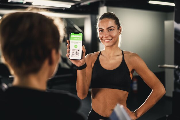 Eine muskulöse junge Frau betritt ein Fitnessstudio und zeigt dem Trainer ihren COVID-Impfpass auf einem Smartphone.