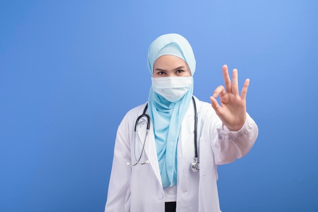 Eine muslimische Ärztin mit Hijab, die eine chirurgische Maske über blauem Hintergrundstudio trägt.