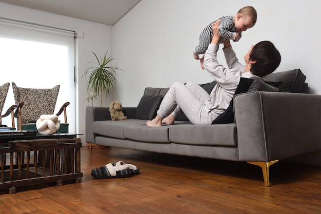 Eine Mutter, die mit dem Baby auf dem Sofa zu Hause spielt, die mit Pyjama