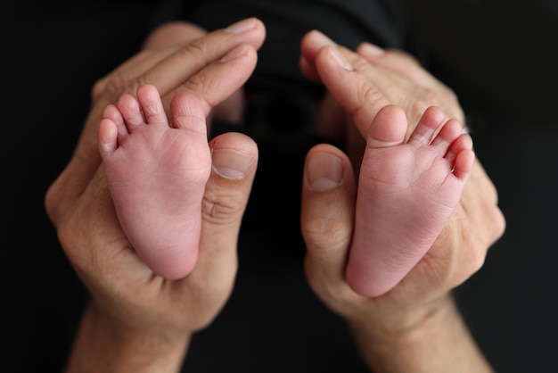 Eine Mutter hält die Füße eines neugeborenen Kindes in einer schwarzen Decke auf schwarzem Hintergrund Die Füße eines Neugeborenen in den Händen der Eltern Studio-Makrofoto Beine, Zehen, Füße und Fersen eines Neugeborenen