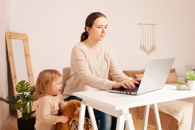 Eine Mutter im Mutterschaftsurlaub sitzt an einem Laptop und arbeitet ein Kind, das hysterisch schreit und ablenkt