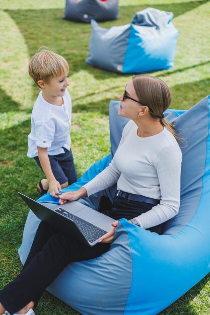 Eine Mutter mit ihrem Sohn auf einer grünen Wiese eine Mutter im Mutterschaftsurlaub arbeitet an einem Laptop im Park ihr Sohn spielt neben ihr