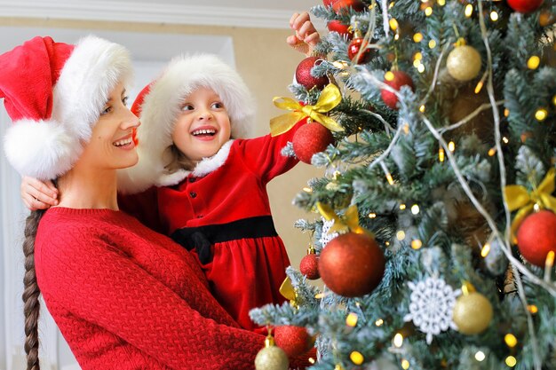 Eine Mutter und ihr Kind in Weihnachtsmützen schmücken den Weihnachtsbaum mit Kugeln an Heiligabend