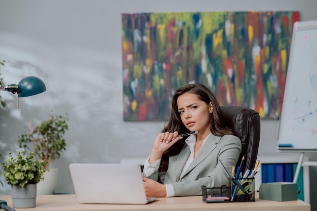 Eine nachdenkliche, arbeitsorientierte Frau sitzt in einem bequemen Ledersessel am Schreibtisch vor einem Laptop