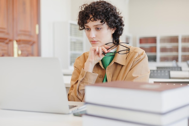 Eine nachdenkliche Frau, die in einer Bibliothek arbeitet, sieht sich ein Bildungs-Webinar im Internet an, ist gelangweilt und arbeitet hinter einem Laptop mit