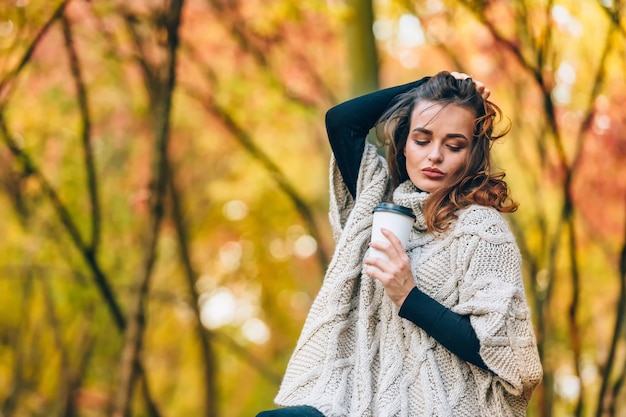 Eine nachdenkliche Frau steht mit einer Tasse Kaffee im Park. Lockiges Haar und schönes Make-up. Hintergrund des Herbstparks. Nahaufnahme