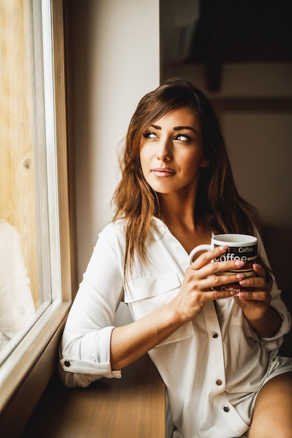 Eine nachdenkliche Frau trinkt Morgenkaffee und schaut durch das Fenster.