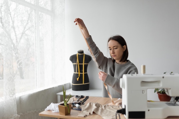 Eine Näherin im Hintergrund ihrer Arbeitswerkstatt ist mit dem Nähen von Kleidung beschäftigt Misst ein Stück Stoff mit einem flexiblen Meter