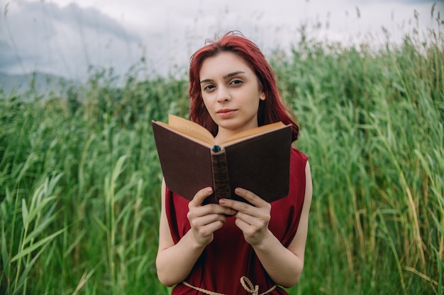 Eine Nahaufnahme des armenischen Mädchens des roten Haares, das Buch in der Natur hält und liest und Kamera betrachtet