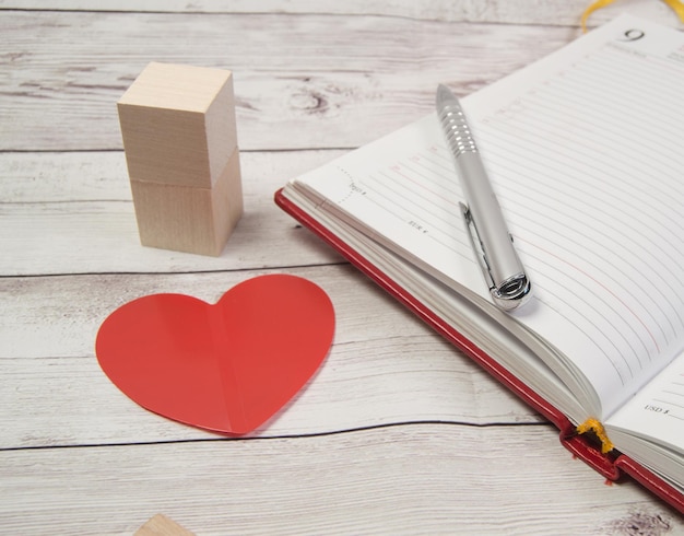 Eine Nahaufnahme eines offenen Tagebuchs für geschäftliche Erinnerungen am Valentinstag mit einem roten Herzen und einem Stift. Holzklötze auf einem Holztischhintergrund