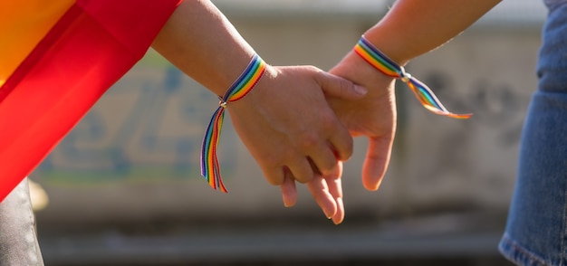 Eine Nahaufnahme von zwei jungen kaukasischen Frauen, die im Freien Händchen mit LGBT-Stolz-Armbändern halten