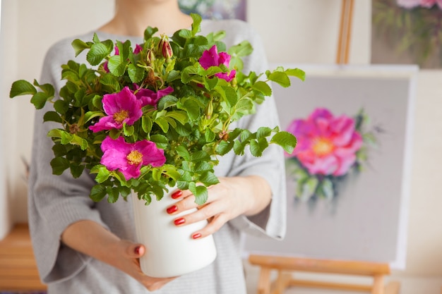 Eine nicht identifizierte verschwommene junge Frau hält eine Vase in ihren Händen mit einer schönen rosa blühenden Hagebutte. Das Konzept der Innendekoration und der Zucht von Zimmerpflanzen