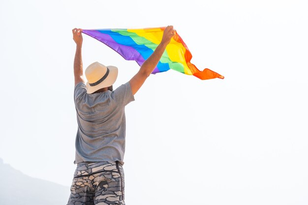 Eine nicht wiederzuerkennende schwule Person in einem grauen T-Shirt und einem weißen Hut, die die LGBT-Flagge an einem bewölkten Himmel schwenkt, Symbol für Homosexualität