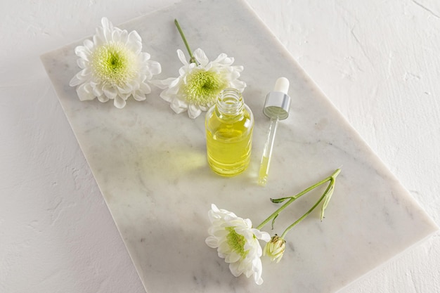 Eine offene Flasche mit kosmetischem Naturheilmittel und eine gefüllte Pipette auf einem weißen Marmorpodium und einem weißen Hintergrund mit Frühlingsblumen-Selbstpflege