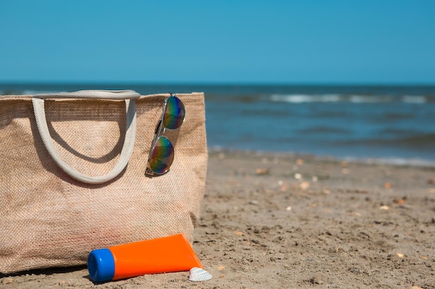 Eine orangefarbene Röhre mit verspotteter Sonnenschutz-Sonnenbrille und eine Strandtasche stehen auf dem Sand in der Nähe des Meeres und schwingen von der Brise UV-Schutz-Strandurlaubsreise